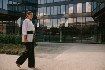 A professional woman confidently walking through an urban landscape while carrying a laptop