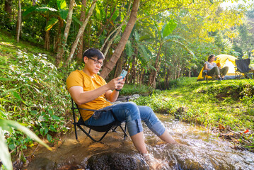 Asian man enjoy outdoor active lifestyle travel nature hiking and camping on summer vacation. Businessman using smartphone remote working corporate business financial at camp in tropical forest.