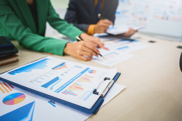 Asian business team engages in a collaborative meeting, financial charts at a desk. Dressed in formal suits, they brainstorm creative ideas to enhance company performance and team cohesion.