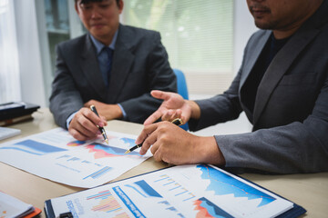 Asian business team engages in a collaborative meeting, financial charts at a desk. Dressed in formal suits, they brainstorm creative ideas to enhance company performance and team cohesion.