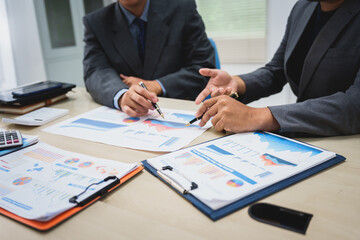 Asian business team engages in a collaborative meeting, financial charts at a desk. Dressed in formal suits, they brainstorm creative ideas to enhance company performance and team cohesion.