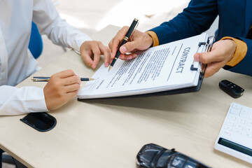 Fototapeta premium A middle-aged Asian male seller in a formal suit assists a woman buyer at a desk. They finalize a vehicle lease, reviewing financial charts and signing contract agreements with a car key.