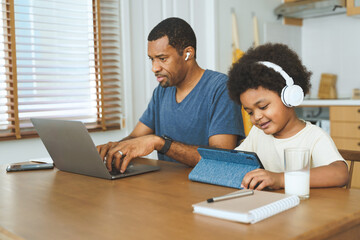 Obraz premium Black African American Father and child using laptop and tablet with headphones at home kitchen table. Casual home lifestyle. Family bonding and home learning concept.