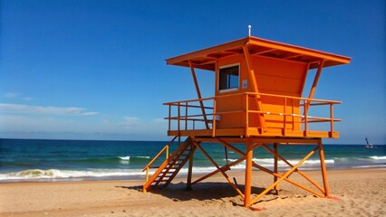 An orange lifeguard tower standing tall on a sandy beach, with the ocean stretching out to the horizon under a clear blue sky.