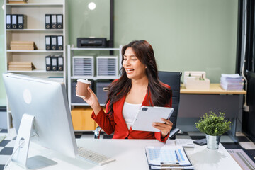 An Asian businesswoman sits at her desk, working efficiently on a computer and tablet. She smiles confidently, displaying her expertise as a financial analyst. Her work brings her happiness