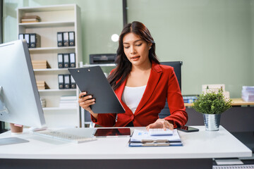 An Asian businesswoman sits at her desk, working efficiently on a computer and tablet. She smiles confidently, displaying her expertise as a financial analyst. Her work brings her happiness