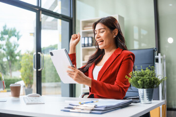 An Asian businesswoman sits at her desk, working efficiently on a computer and tablet. She smiles confidently, displaying her expertise as a financial analyst. Her work brings her happiness