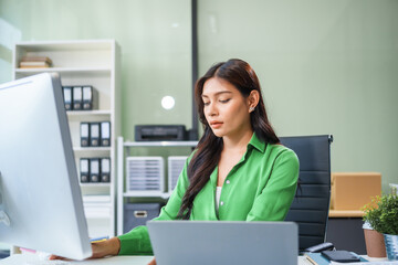 An Asian businesswoman sits at the table in her office, working as an accountant. She uses a computer with a smile, feeling happy and content with her work.