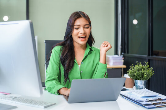 An Asian businesswoman sits at the table in her office, working as an accountant. She uses a computer with a smile, feeling happy and content with her work.