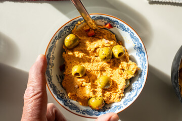 Hummus in a bowl with olives, a serving spoon, and a hand.