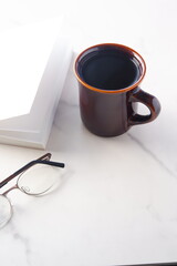 白いテーブルに置かれた一冊の本とメガネ、コーヒーの入ったマグカップで、コーヒーを飲みながら読書を楽しむイメージ

