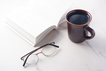 白いテーブルに置かれた一冊の本とメガネ、コーヒーの入ったマグカップで、コーヒーを飲みながら読書を楽しむイメージ
