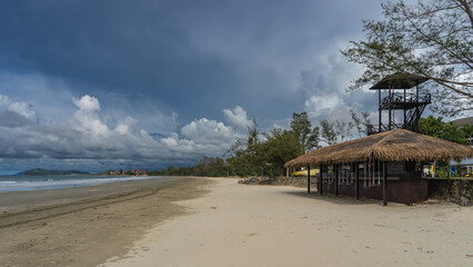 A bar on a sandy beach. Thatched roof. Swing seats are suspended at the bar counter. A lifeguard observation tower against a blue sky, clouds. The waves of the turquoise ocean are foaming. Malaysia