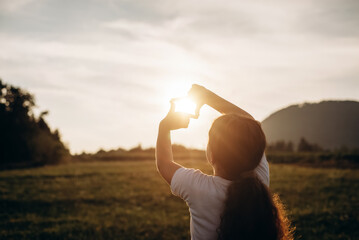 Preteen girl making frame gesture at amazing sunset in beautiful summer park. Concept of seeing world as different. Business planning. Little child shows fingers frame symbol at sunrise