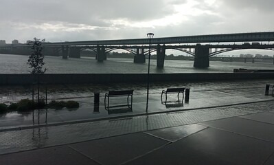 A serene cityscape with a long bridge over a wide river under a cloudy sky. Silhouetted bridge and...