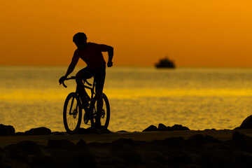 Fototapeta premium A man rides a bike atop a sandy beach, nearby lies a tranquil body of water In the distance, a boat floats