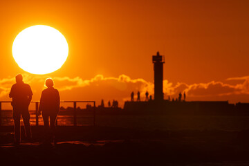  A few individuals stand adjacent to one another by a lighthouse as the sun sets, casting a golden...