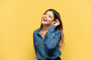 Young Russian girl isolated on yellow background listening to something by putting hand on the ear