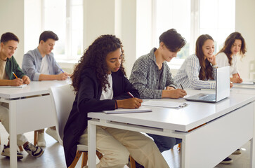 Side view of a group of diverse young high school people students sitting at the desk in a row and writing test during a college lesson in the classroom. Education and knowledge concept.