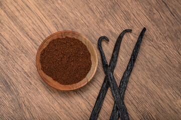  Vanilla pods lie on a wooden kitchen counter next to ground dried vanilla spice in a wooden bowl