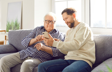 Happy adult son helps elderly father understand the phone and modern technology, sitting together...