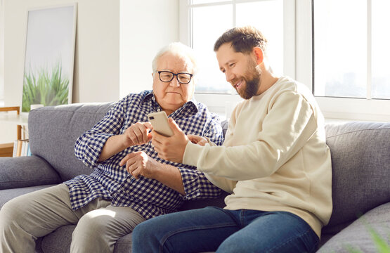 Happy adult son helps elderly father understand the phone and modern technology, sitting together on the sofa at home. Child learning old parent about the internet and online tools.