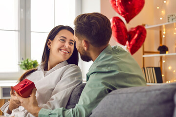 Obraz premium Young couple in love sitting on the sofa at home and exchanging romantic gifts. Man gives a present to a happy, smiling beautiful young woman on St Valentine's Day. Love and romance concept