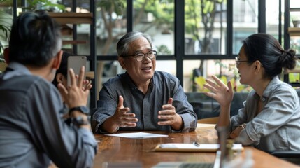 A group of three colleagues engaged in an animated discussion around a wooden table with large windows and greenery in the background.