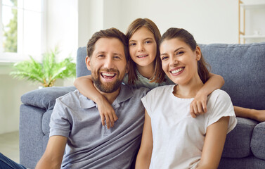 Happy family at home. Portrait of cheerful, smiling parents together with their child. Loving mother, father and little daughter sitting on floor by couch at home, looking at camera and smiling