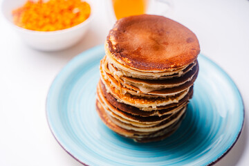 Pancakes stacked on a turquoise plate on a white background with sea buckthorn and tea.