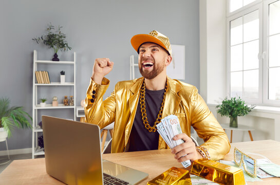 Successful excited young funny man in golden suit sitting at desk with gold bar, building online business using laptop, holding money cash of dollar bills celebrating success or win a lottery.