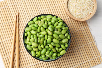 Fresh edamame soybeans in bowl, chopsticks and sesame seeds on white wooden table, top view