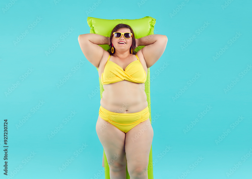 Wall mural Happy funny plus size woman in sunglasses and in summer yellow bikini lying on inflatable mattress isolated on a studio blue background. Fat girl going to swim on a beach. Vacation concept.