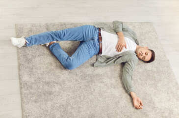  Man lies unconscious on grey carpet in home, possibly due to accident or health problems. Top view of young man in casual clothes lying with his eyes closed on floor holding his hand over his heart.