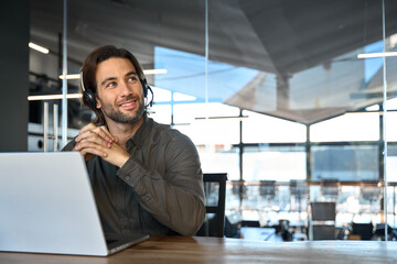 Happy male customer support service tech operator sitting at work desk looking away. Young...