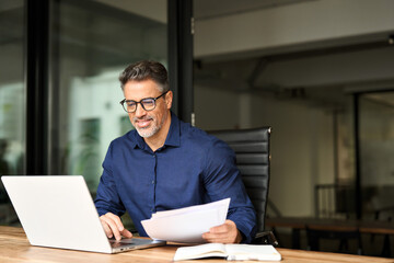 Happy busy older professional businessman entrepreneur using laptop holding bill corporate document...