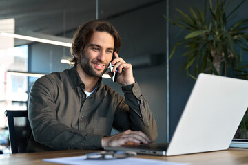 Happy young business man sales manager talking on mobile phone working on laptop in office. Smiling professional businessman making call on cell using computer communicating with client at work.