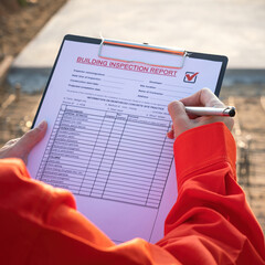 A project engineer in orange coverall uniform is using a pen to checking on building quality checklist to inspecting the quality, with background of construction work site. Close-up.