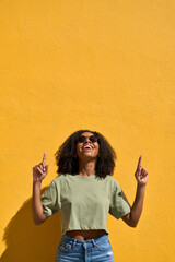 Young happy African gen z girl in sunglasses standing at yellow wall pointing up at copy space. Smiling Black woman presenting promo on summer day advertising shopping sale offers, vertical photo.