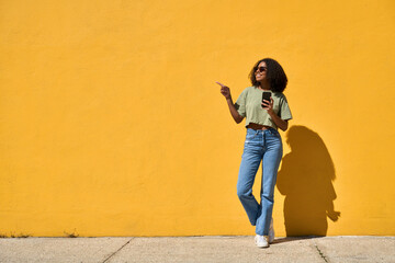 Obraz premium Happy African American gen z girl wearing sunglasses standing at yellow wall pointing at copy space holding smartphone. Smiling young Black woman using mobile phone advertising summer sale. Full body.