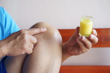 Close up hand holds bottle of herbal yellow balm to apply on his knee. Concept, Health care. First aid. Self care at home, use herbal ointment to relieve pain, injury sprain.             