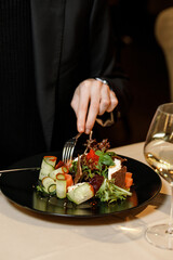 handsome man in a black sweater is eating a delicious salad with juicy meat, fresh vegetables, fragrant herbs, and crispy croutons, served on a black plate in a cozy, charming restaurant