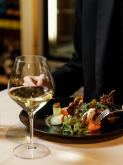 handsome man in a black sweater is eating a delicious salad with juicy meat, fresh vegetables, fragrant herbs, and crispy croutons, served on a black plate in a cozy, charming restaurant