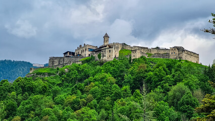 Obraz premium Panoramic view Landskron Castle in Villach, Carinthia, Austria. Majestic fortress perched atop verdant forest covered hill. Imposing structure with fortified walls, towers and prominent church spire