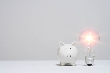 White piggy bank and glowing light bulb side by side on a plain background, symbolizing financial...