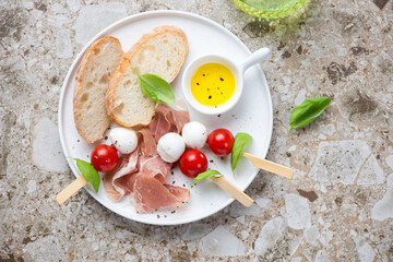 Prosciutto, caprese skewers and ciabatta on a white plate, top view on a light-brown granite background, horizontal shot