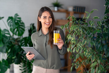 A cheerful Caucasian woman sips on fresh juice while expertly navigating a digital tablet,...