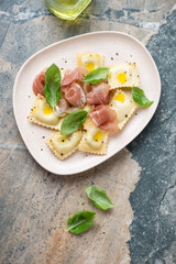 Italian ravioli served with prosciutto and basil, above view on a beige and grey granite background, vertical shot with space