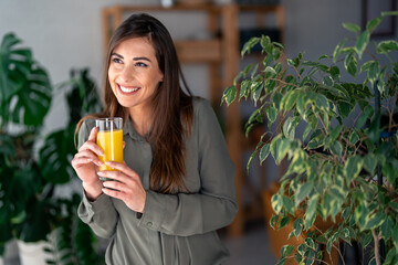 A joyful Caucasian lady sips orange juice, dressed casually for her professional environment, surrounded by greenery enhancing the freshness of a contemporary workspace.