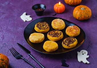 Halloween dessert, pumpkin cheesecakes with chocolate spider web decor on black ceramic plate on purple concrete background.
